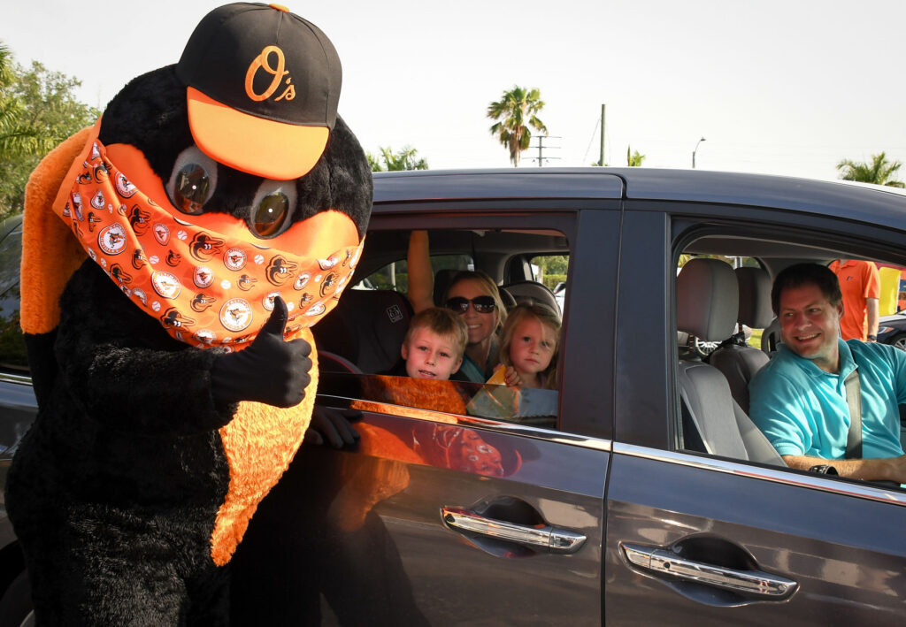 A person in an Orioles mascot costume gives a thumbs-up beside a car with two children and two adults inside, all smiling at the camera.