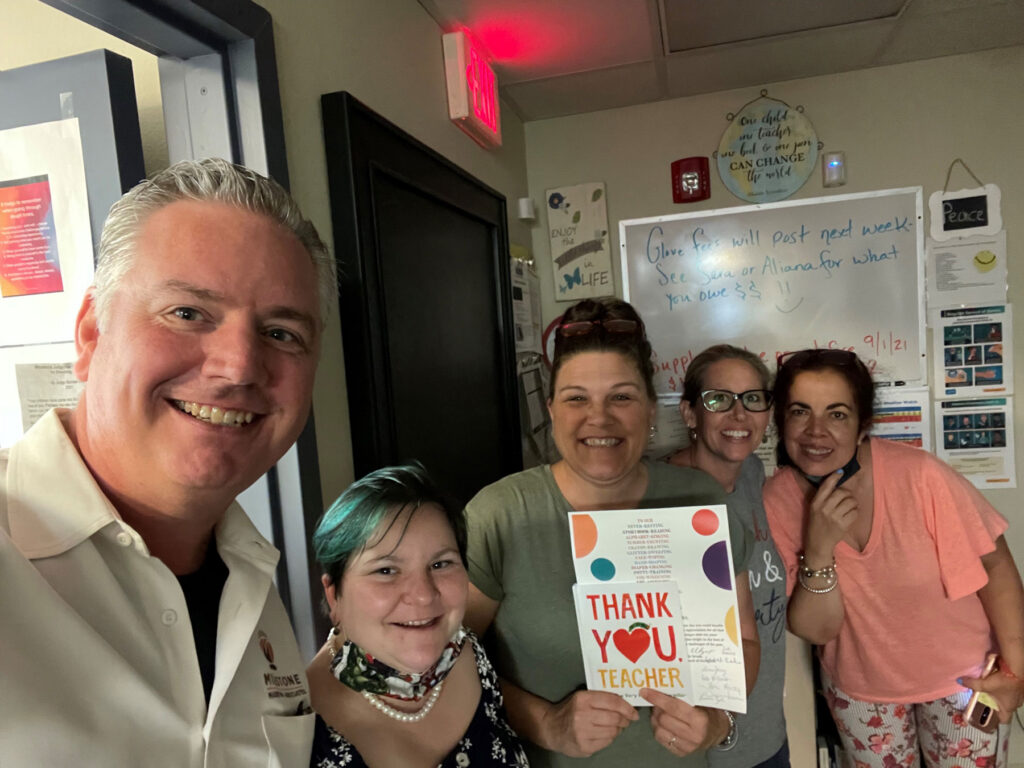 Five people stand in a classroom, smiling at the camera. One holds a colorful "Thank You Teacher" sign. The background features posters and notes on the wall.