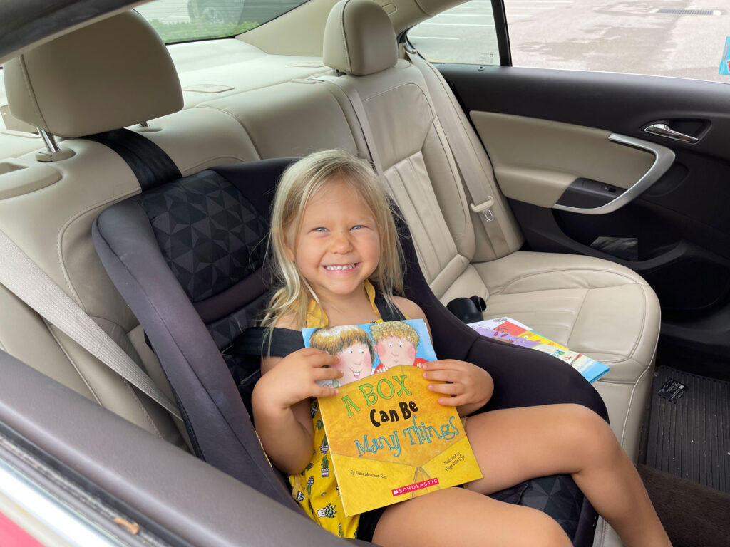 A young child sits in a car seat in the back of a car, smiling and holding a picture book titled "A Box Can Be Many Things.