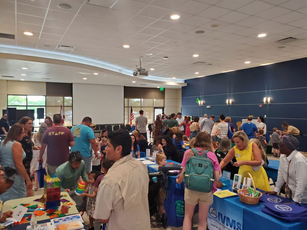 Crowd of adults and children visit various information tables in a large, well-lit room during a community or school event.