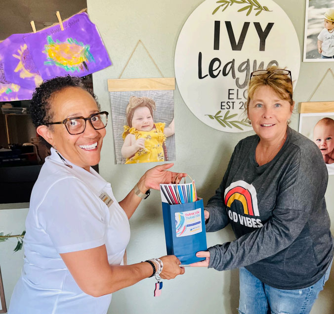 Two women stand indoors, holding a container filled with colored folders. Children's artwork and photos hang on the wall behind them, with a sign reading "IVY League ELC EST 2019.