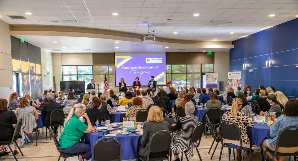 A group of people attend an Educator Business Breakfast event in a large room, with a panel of speakers at the front and a screen displaying "Business Breakfast of Champions.